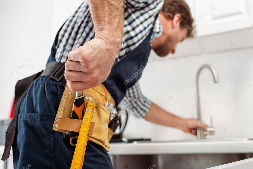 Selective focus of plumber taking wrench from tool belt while fixing faucet in kitchen 