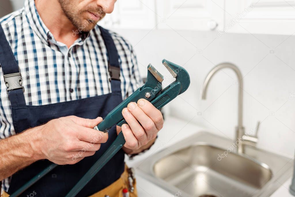 Cropped view of workman in overalls holding pipe wrench in kitchen 