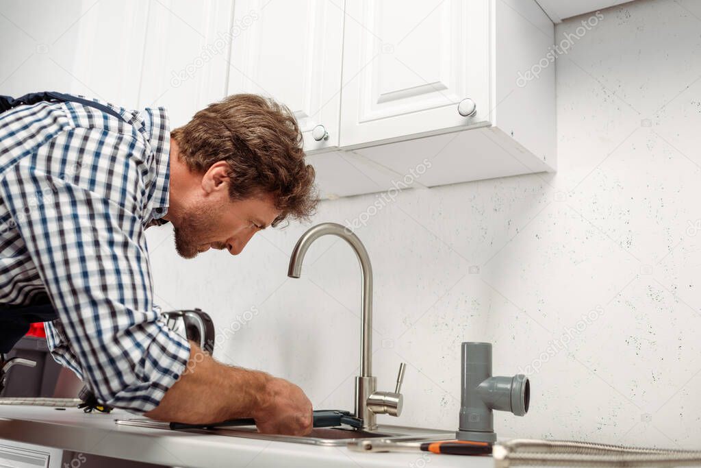 Side view of plumber holding pipe wrench while repairing kitchen faucet near tools on worktop 