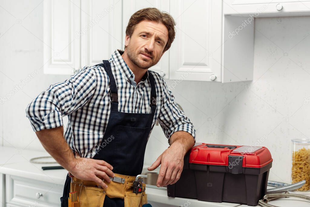 Handsome workman in tool belt looking at camera near toolbox on kitchen worktop 