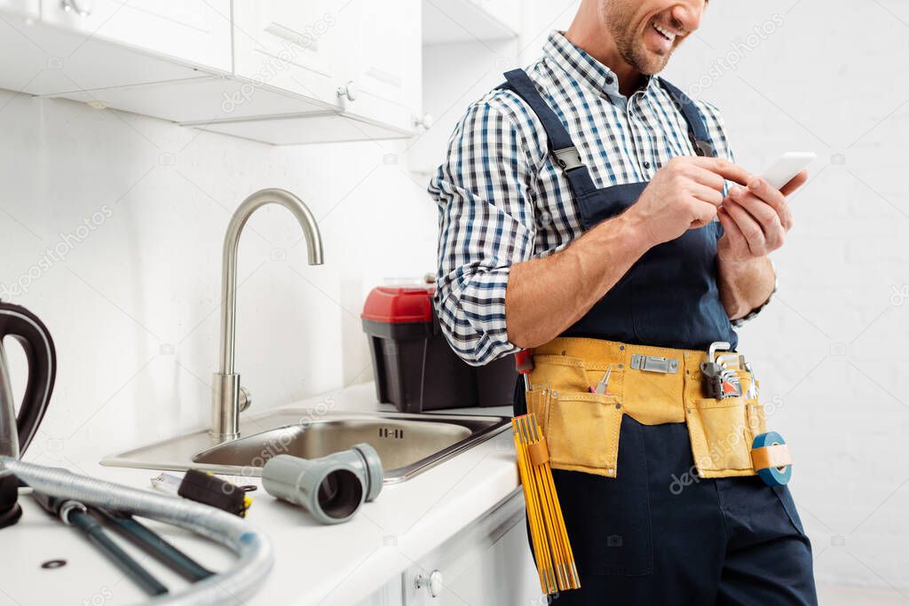 Cropped view of smiling plumber using smartphone near tools on kitchen worktop 