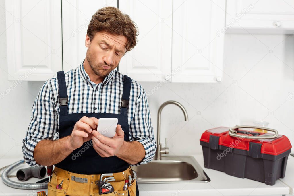 Pensive plumber using smartphone near toolbox and sink in kitchen 