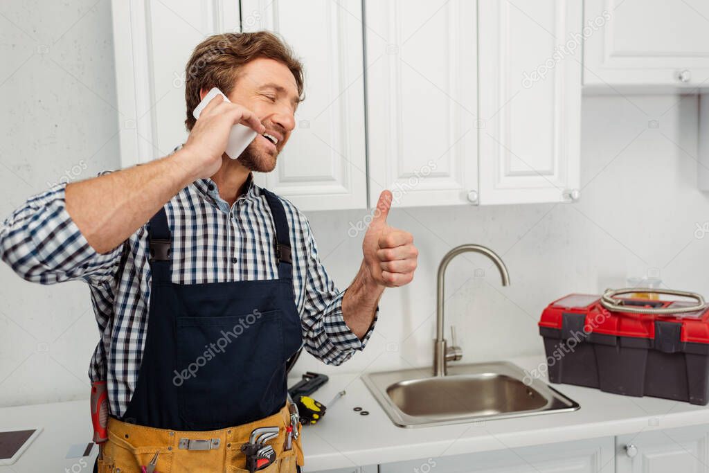 Smiling plumber showing like gesture and talking on smartphone near kitchen sink 