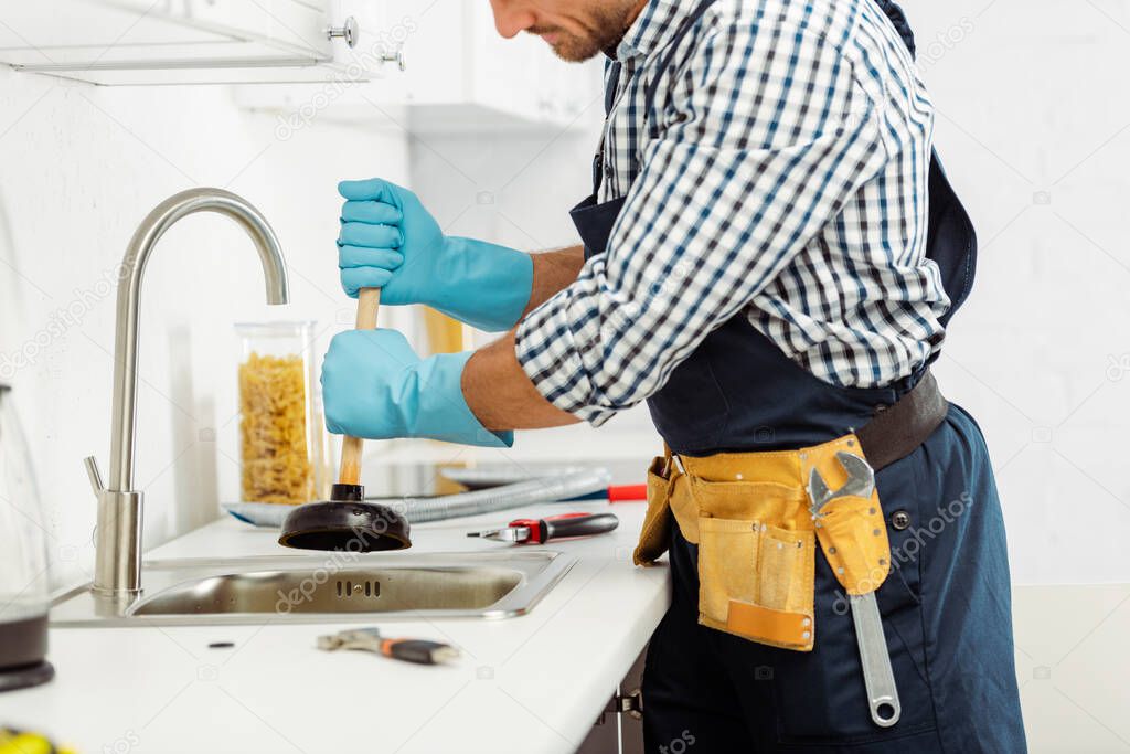 Cropped view of plumber in tool belt and rubber gloves holding plunger near tools on kitchen worktop 