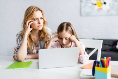 Evde çevrimiçi eğitim sırasında dizüstü bilgisayara bakan düşünceli anne ve çocuğun seçici odağı 