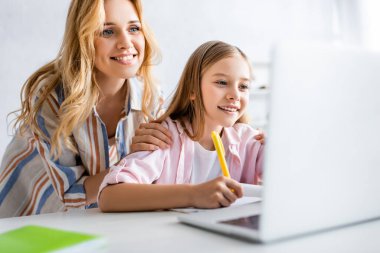 Elektronik öğrenme sırasında neşeli kızı kucaklayan gülümseyen kadının seçici odağı 