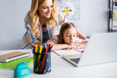 Gülümseyen bir kadının evde online eğitim sırasında üzgün çocuğa sarılmasının seçici odağı. 