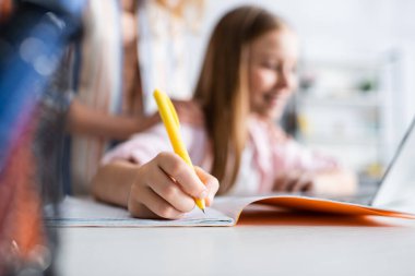 Selective focus of kid writing on notebook during electronic learning near mother   clipart