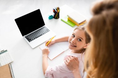 Overhead view of smiling kid looking at mother while writing on notebook near laptop on table  clipart