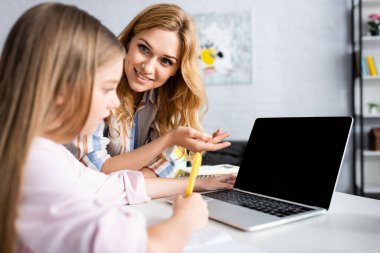 Çevrimiçi eğitim sırasında, kızın yanındaki dizüstü bilgisayarı işaret eden gülümseyen kadının seçici odağı 
