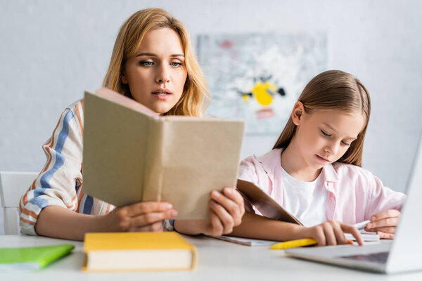 Selective focus of woman reading book while helping daughter during online education at home 