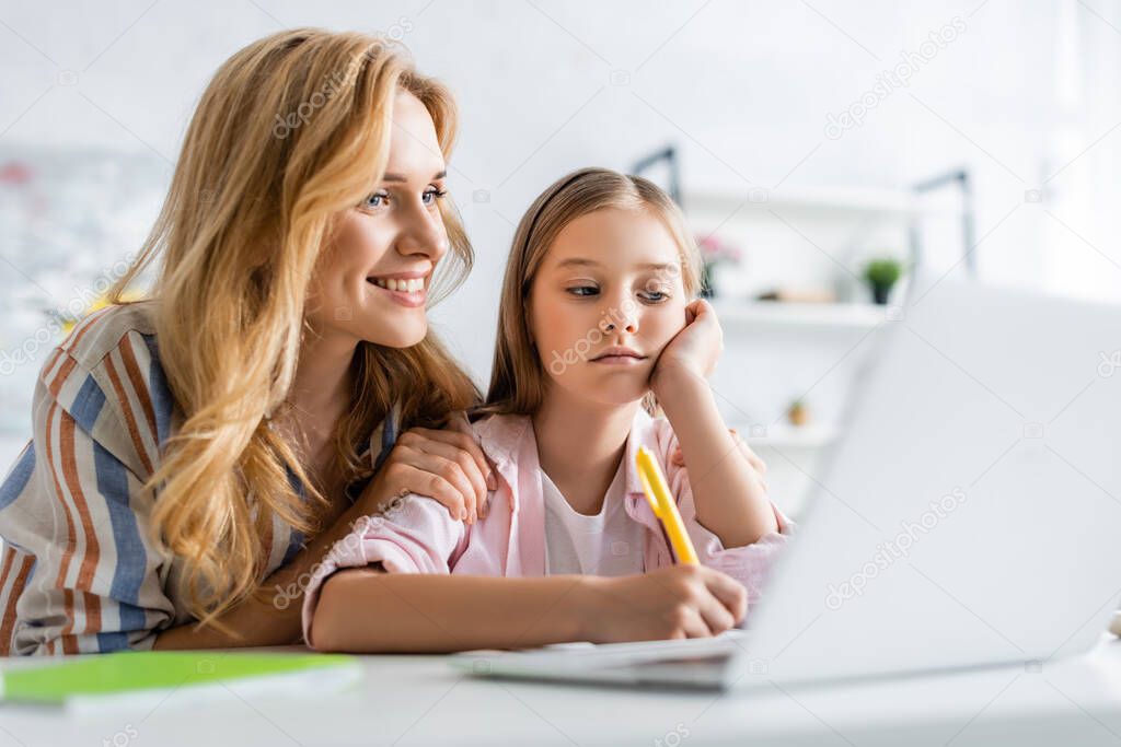 Selective focus of smiling mother sitting near daughter writing on notebook near laptop on table 