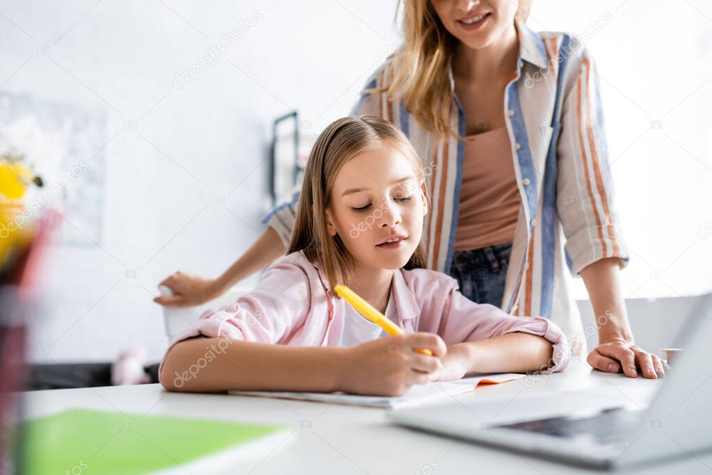 Selective focus of smiling mother standing near daughter writing on notebook near laptop on table 