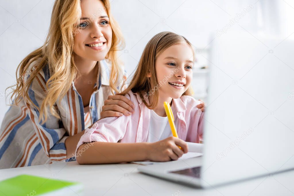 Selective focus of smiling woman embracing cheerful daughter during electronic learning 
