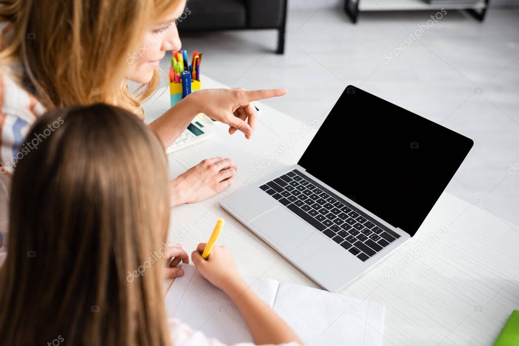 Selective focus of woman pointing with finger at laptop near kid writing on notebook during electronic learning at home 