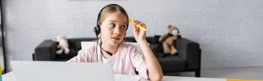 Panoramic orientation on thoughtful kid in headset holding pen during electronic learning  clipart