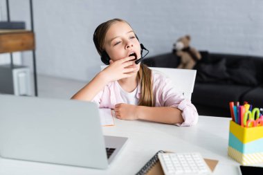 Evde elektronik öğrenim sırasında esneyen kulaklıklı uykulu çocuğun seçici odağı 