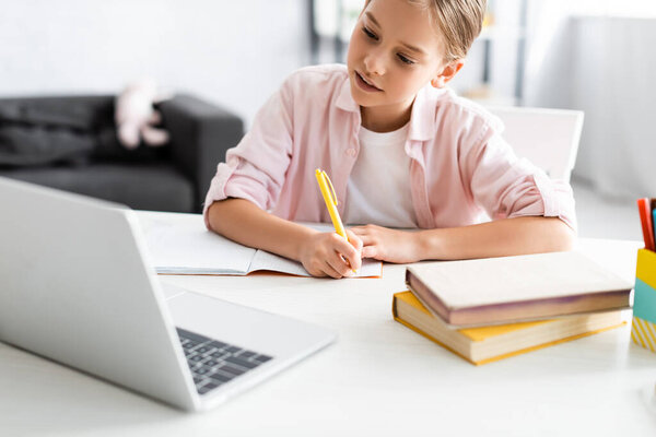 Selective focus of cute child writing on notebook near books and laptop during electronic education 