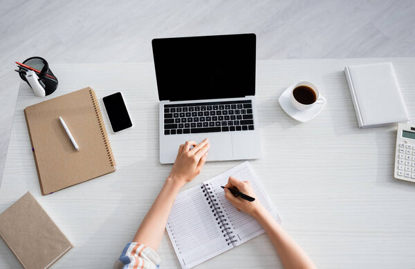 Cropped view of woman writing in notebook and working with laptop