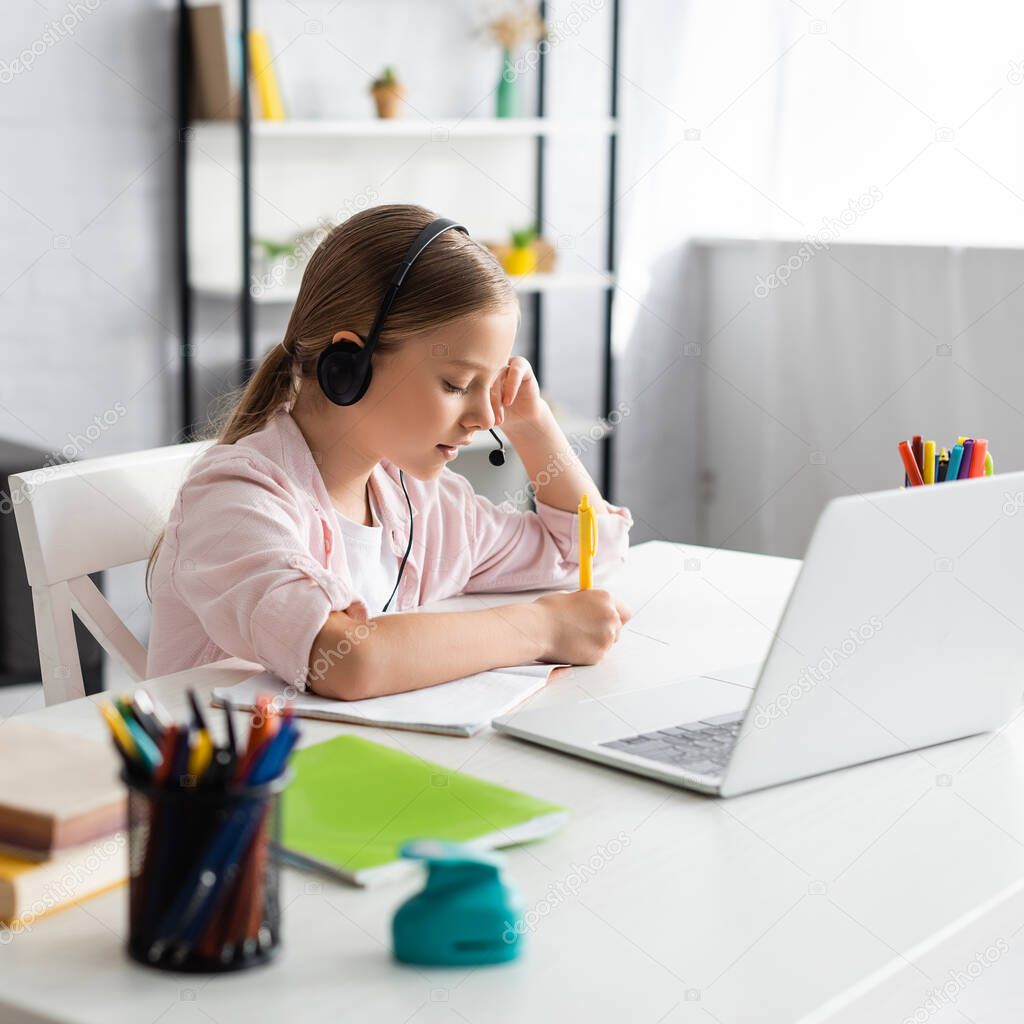Selective focus of kid in headset writing on notebook near laptop and stationery on table 