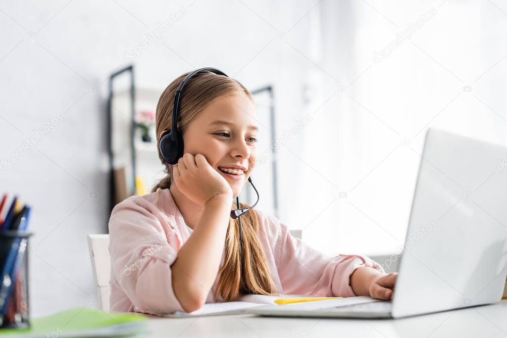 Selective focus of smiling kid using headset and laptop at table 