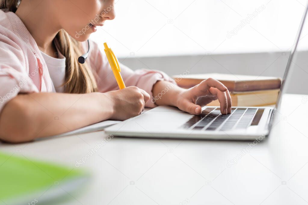 Cropped view of kid in headset writing on notebook and using laptop at home 