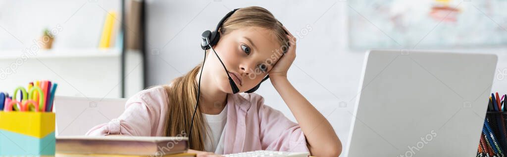 Panoramic shot of tired kid in headset looking at laptop near stationery