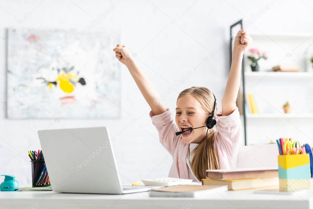 Selective focus of cheerful kid showing yeah gesture during online education near stationery on table 