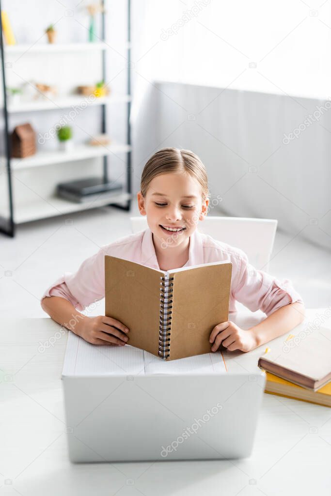 Selective focus of smiling kid holding notebook near laptop and books on table 