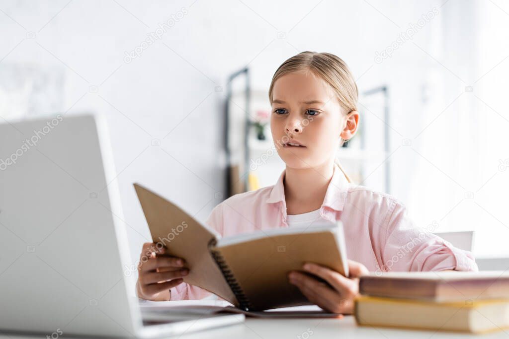 Selective focus of concentrated kid looking at laptop while holding notebook at table 