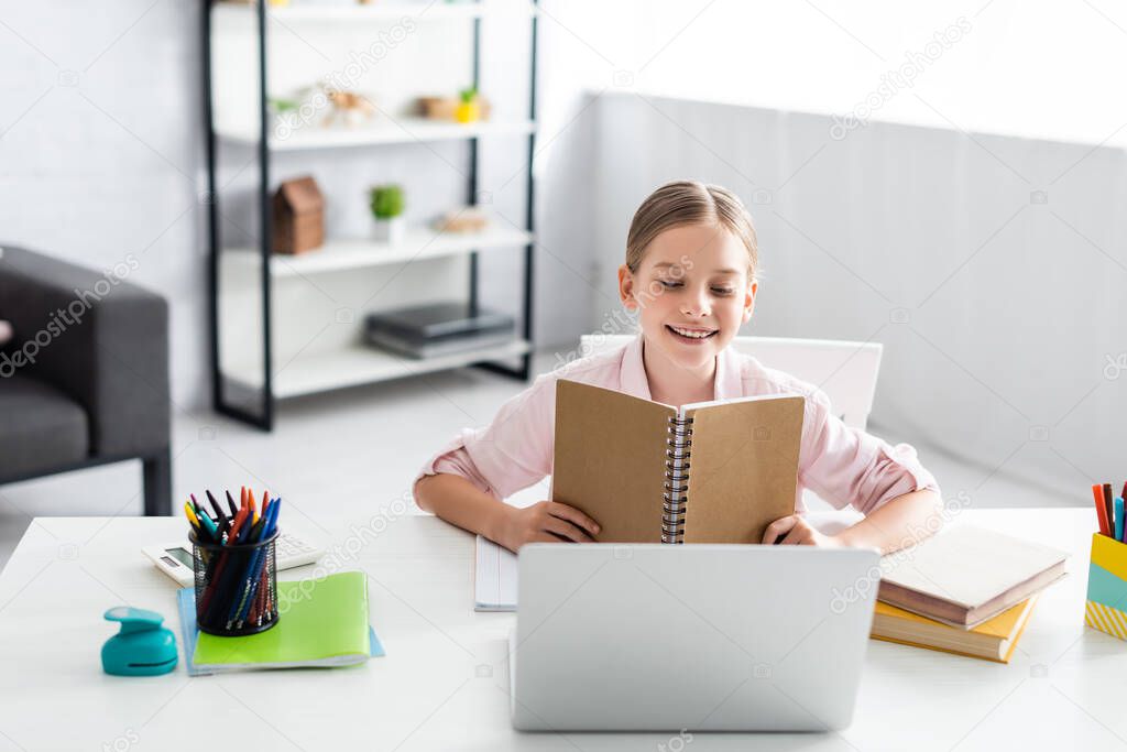 Selective focus of smiling kid holding notebook near laptop and books on table at home 