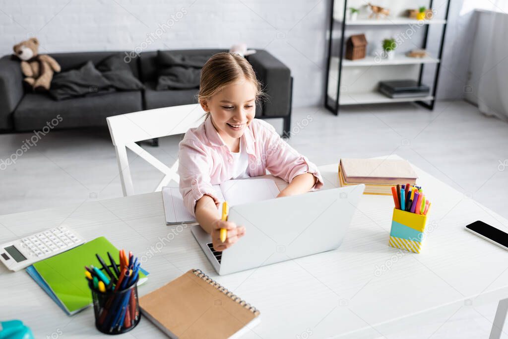 Selective focus of smiling child holding pen and using laptop near books on table 