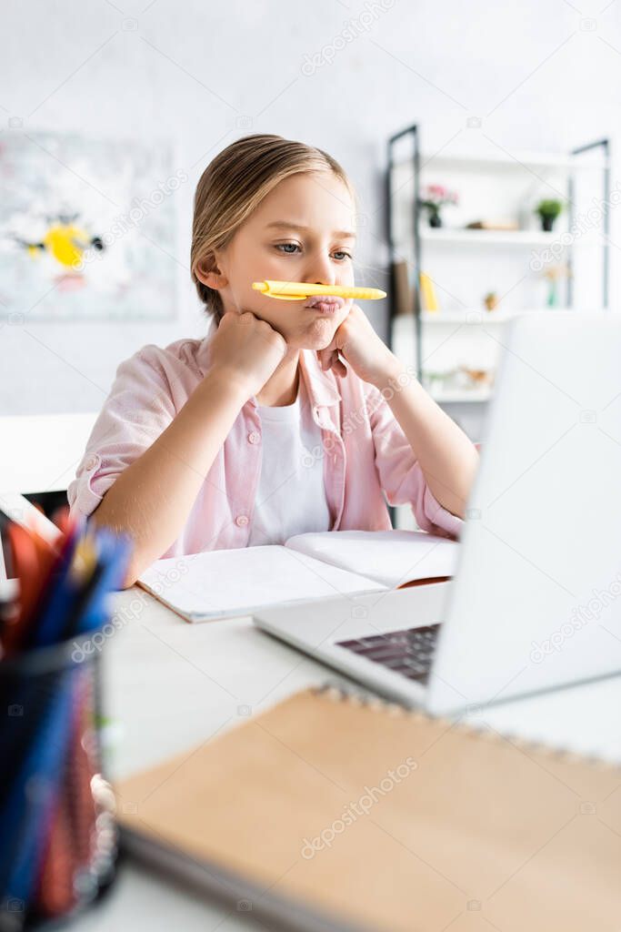 Selective focus of kid holding pen near lips during electronic learning at home 