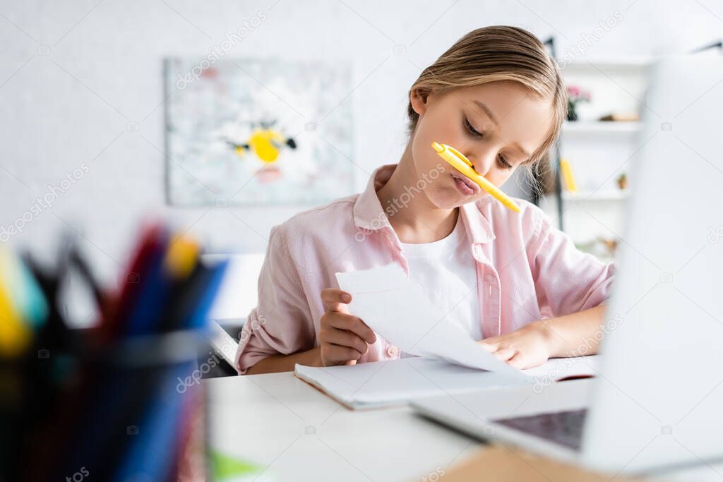 Selective focus of kid with pen near mouth looking at notebook near laptop 