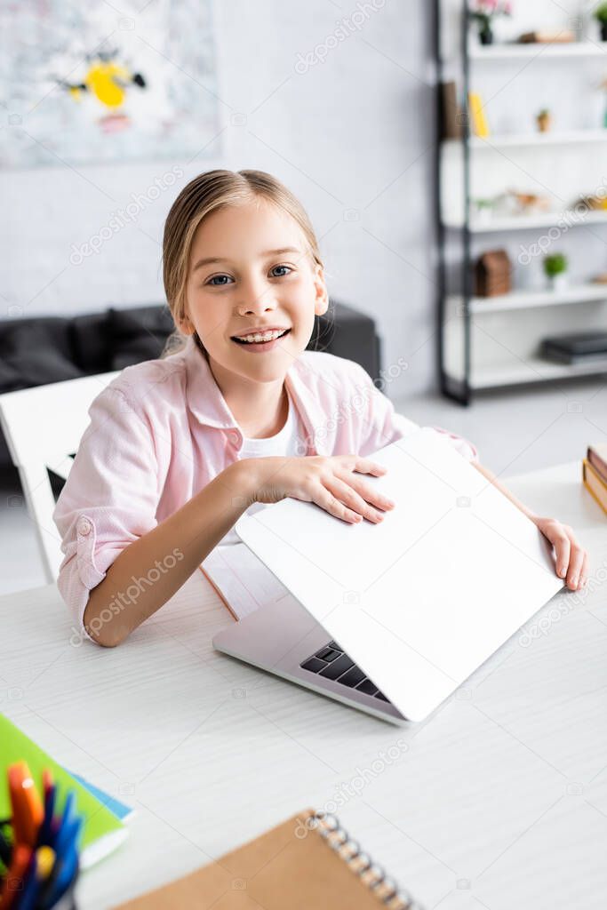 Selective focus of smiling kid looking at camera while holding laptop near stationery on table 