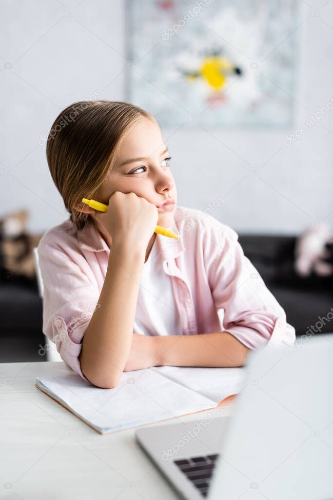 Selective focus of dreamy kid with pen looking away near laptop and copy book on table