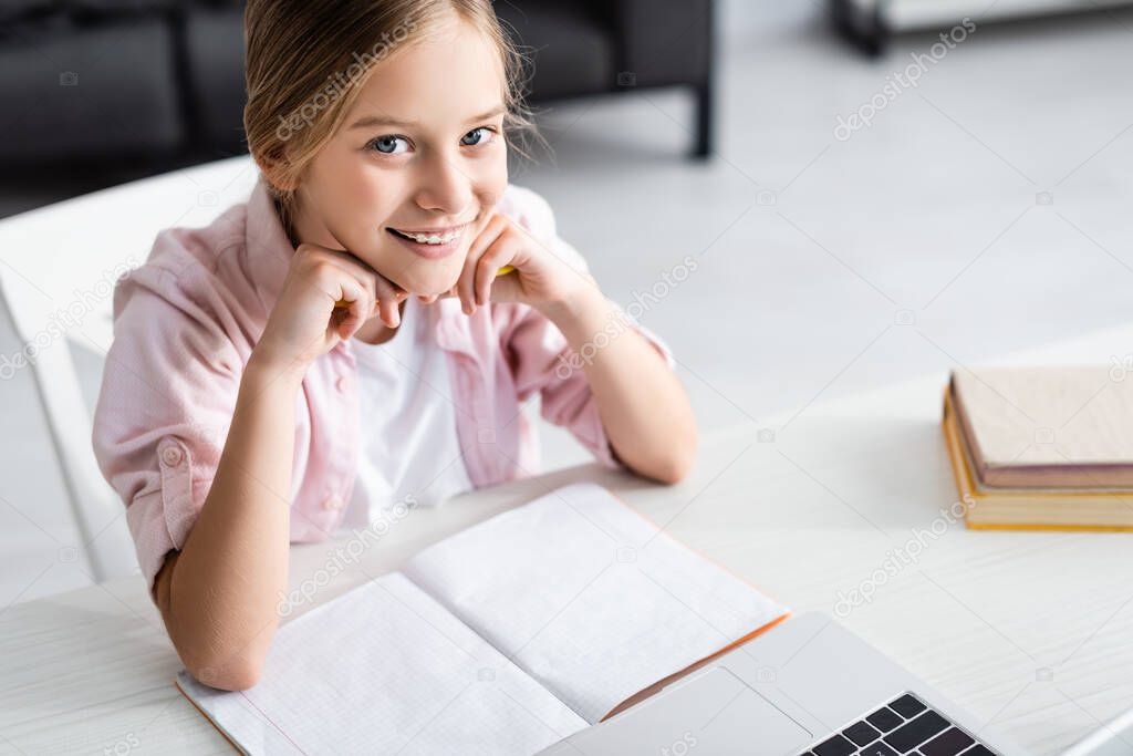 High angle view of cute child smiling at camera near laptop and copy book on table 