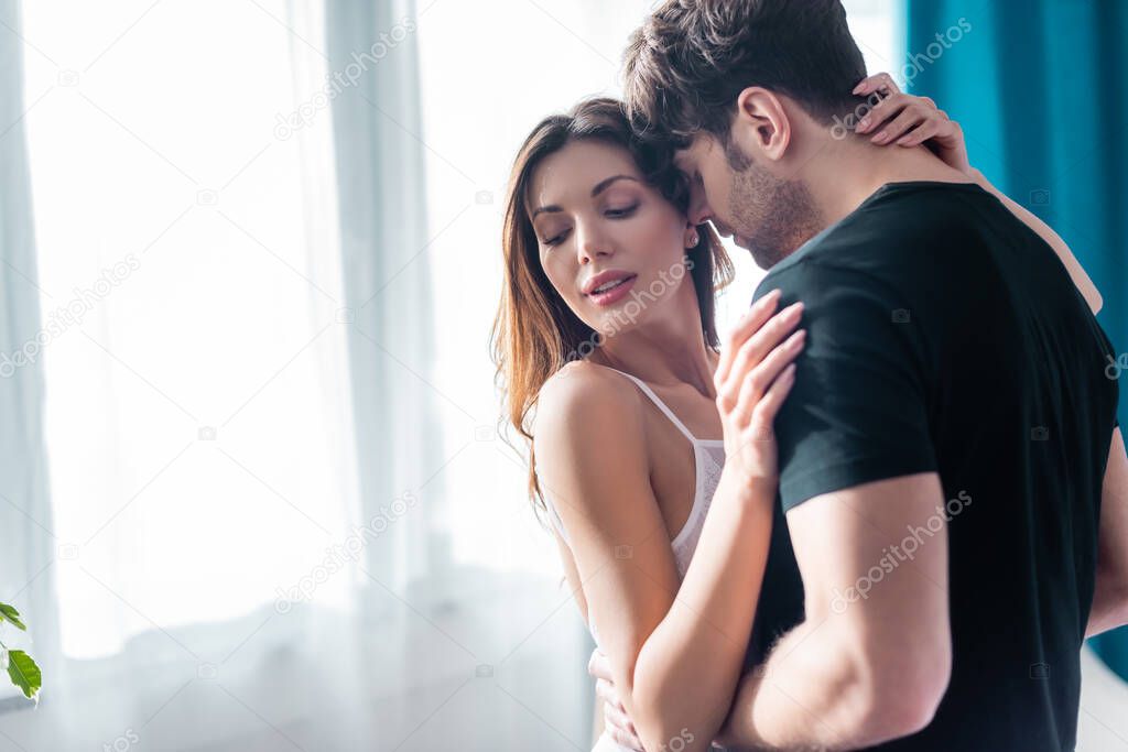 Selective focus of young man embracing attractive girl near window at home 