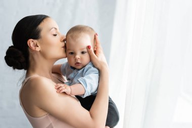 Anne kollarını tutuyor ve bebek kıyafetleri içinde tatlı bir çocuğu öpüyor. 