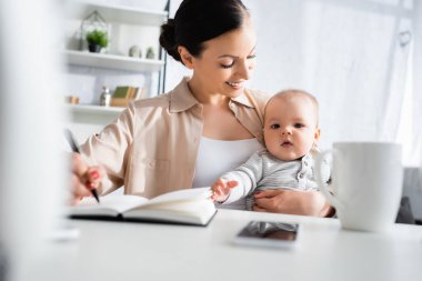 Seçici odak noktası, elinde kalem tutan mutlu anne ve akıllı telefonun yanındaki küçük tatlı çocuğa bakıyor. 