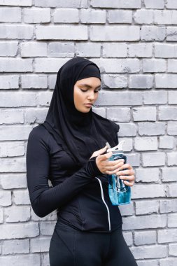 tesettürlü genç müslüman sporcu kadın dışarıda suyla birlikte spor şişesine bakıyor. 
