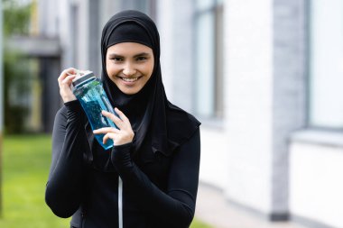 tesettürlü neşeli müslüman sporcu kadın suyla spor şişesini tutuyor ve kameraya bakıyor.
