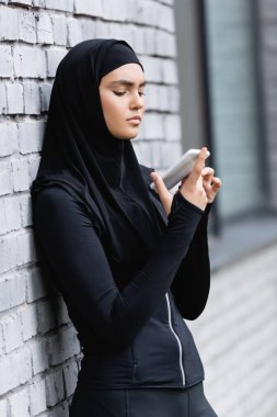 Genç Müslüman kadın akıllı telefon kullanıyor ve tuğla duvarın yanında duruyor.