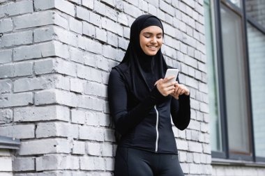 Mutlu Müslüman kadın akıllı telefon kullanıyor ve tuğla duvarın yanında duruyor. 