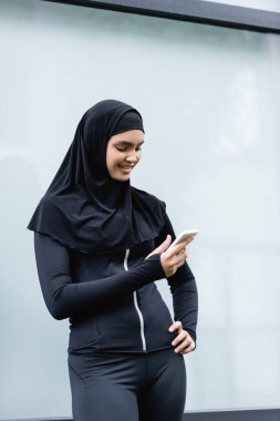 happy muslim woman using smartphone and standing with hand on hip  clipart