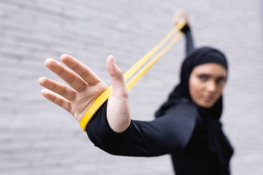 selective focus of arabian sportswoman in hijab exercising with resistance band near brick wall clipart