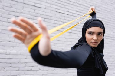 selective focus of arabian woman in hijab exercising with resistance band near brick wall clipart
