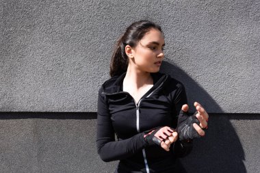 Genç ve çekici bir kadın beton duvarın yanında spor eldivenlerine dokunuyor.