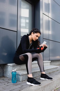 Genç bayan spor şişesinin yanında oturuyor ve akıllı telefon kullanıyor. 