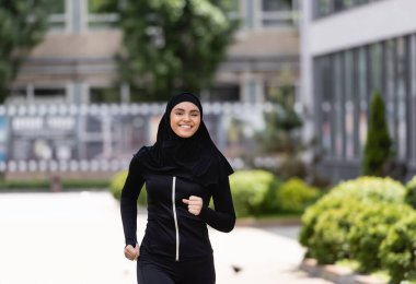 tesettürlü ve spor kıyafetli gülümseyen Arap kız dışarıda koşuyor.